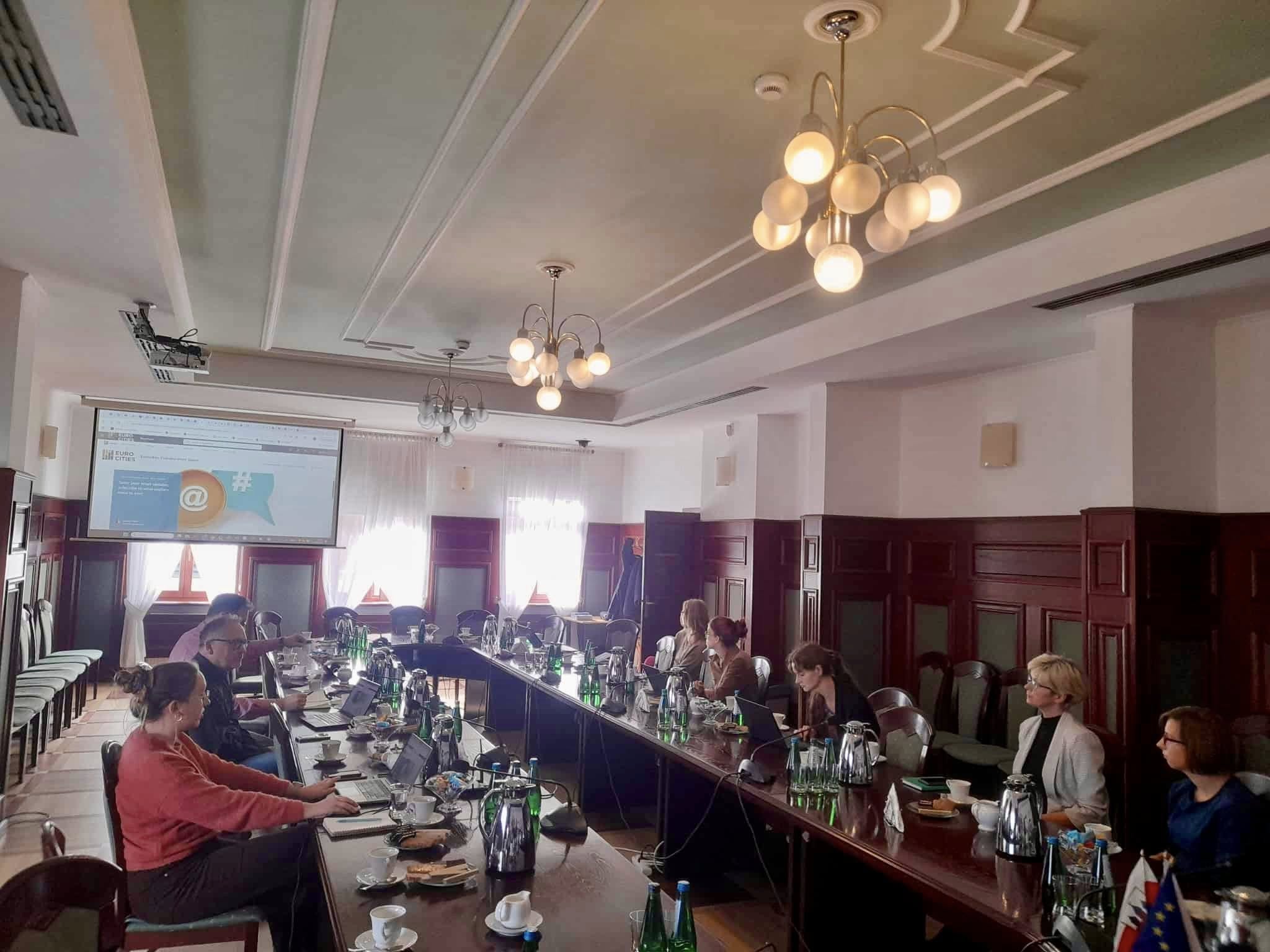 zdjęcie rpzedstawia widok na salę ratuszową, przy owalnym stole siedzą uczestnicy spotkania, przedstawicielka Eurocities pokazuje prezentację